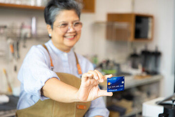 Senior female entrepreneur holding credit card with smile at cafe counter, representing payment...