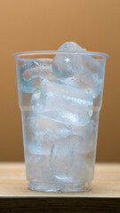 Clear plastic cup filled with transparent ice cubes, placed on a wooden surface against a warm...