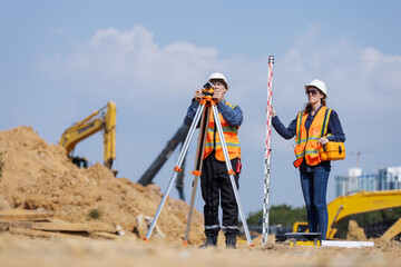 Two construction surveyors in safety gear stand at a job site using a tripod-mounted total station and leveling rod to measure land elevation, essential for site layout and infrastructure planning.
