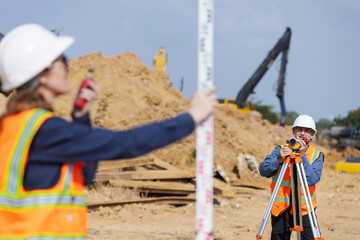 A construction surveyor uses a total station while another holds a leveling rod at a worksite filled with dirt and machinery, capturing accurate land measurements for site planning and layout.