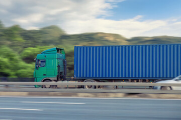 Green truck with blue cargo container moving fast on highway in countryside scene.