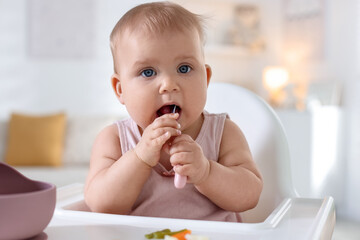 Cute little baby eating with spoon on feeding chair at home