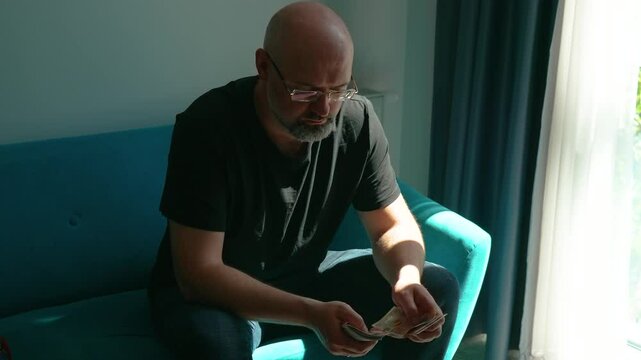 Man counting salary in pound cash. Checking earnings in British currency near window. Handling ten pound notes on sofa in daylight. Managing paper money with thoughtful expression indoors