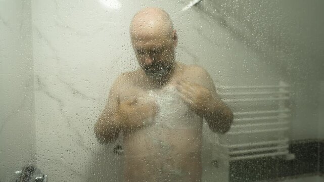 Man washing chest behind steamy shower glass. Rubbing soap on body during morning hygiene. Shower scene with condensation on bathroom door. Daily routine in progress with water droplets blurring view