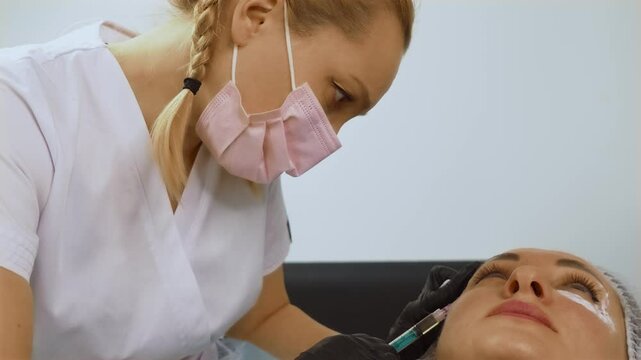 Cosmetologist injecting anti-aging filler under eye area of woman patient during facial treatment. Female aesthetician performing cosmetic procedure with syringe to rejuvenate skin. Specialist