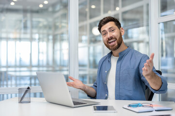 A businessman in a modern office, smiling and gesturing while using a laptop at his desk.