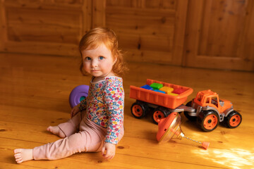 Adorable red-haired toddler girl sitting on a wooden floor in a cozy room, surrounded by bright plastic toys including a toy truck and a spinning top.