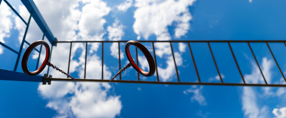 Close-up view of metal gymnastic rings and horizontal ladder bars on a playground, with blue sky and clouds in the background.