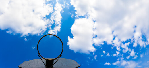 Low angle view of a weathered basketball hoop with no net, framed by a bright blue sky and white clouds.