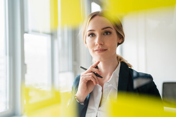 Thoughtful businesswoman with pen looking at adhesive note seen through glass