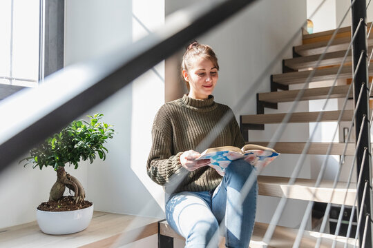Young woman reading book while sitting on staircase at home
