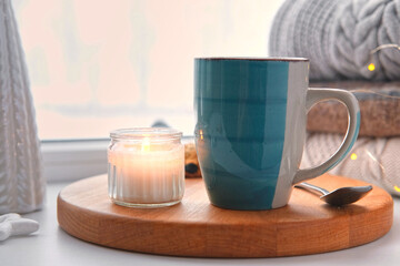 Winter windowsill still life. Blue ceramic cup of hot coffee on window sill. Christmas decorations on the background. Cozy home picture. Warm woolen knitted sweaters, Burn Candle, Cookies. Stock photo