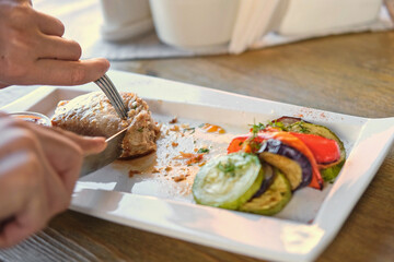 Girl cuts a cooked pork steak served on a white plate with grilled vegetables. Young woman using knife and fork to cutting meat eating in a restaurant. Balanced diet, cooking, culinary. Food concept