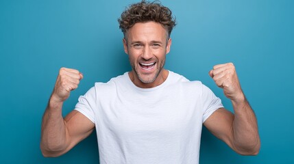 Energetic man with curly hair wearing a white t-shirt, expressing excitement with raised fists against a vibrant blue background, showcasing joy and enthusiasm in a celebratory moment