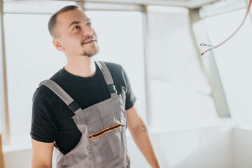 A cheerful electrician wearing gray overalls is assessing the workspace in a bright, modern...
