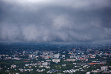 aerial view of chiang mai cityscape under a cloudy, rainy season sky, with urban buildings, a winding road, and green trees partially obscured by mist.
