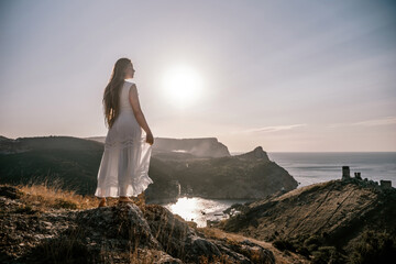 A woman stands on a rocky hill overlooking the ocean. She is wearing a white dress and she is...