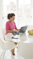 Middle aged woman is sitting at the table and working on a laptop at home.  White colored interior of apartment.