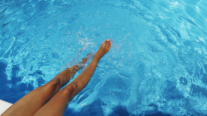 Woman is sitting at the edge of the pool with her legs dangling in the water and shaking the water  and making ripples and small waves. Summer background.