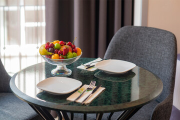 Softly lit secene of colorful fresh fruit bowl on marble tabletop with plates and chairs in the background in front of a window