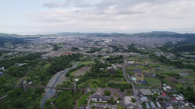 Aerial view of Hadano, Japan, showing lush green farmland, rural houses, and surrounding forested mountains under an overcast sky. A peaceful countryside scene captured by drone.