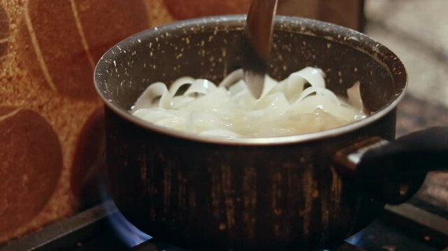 Stirring flat rice noodles known as Kuey Teow, Kuay Tiew, or Hor Fun boiling in a pot on a stove in preparation for cooking stir-fry dish