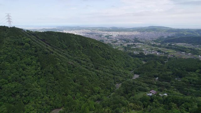 Drone footage of power lines stretching across lush forested hills in Hadano, Japan. A dramatic contrast between nature and infrastructure with a distant cityscape in the background.
