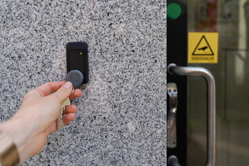 Close-up of a female hand holding a keychain with a key fob near a key reader access device on a building’s exterior wall with door and video surveillance sign. Secure access control technology
