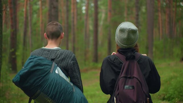 Students walking side by side through forest trail during outdoor exploration, boy carrying sleeping bag and girl adjusting backpack while looking around forest environment surrounded by trees