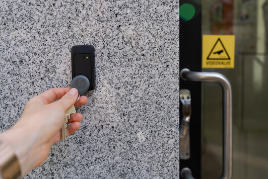 Close-up of a female hand holding a keychain with a key fob near a key reader access device on a building&rsquo;s exterior wall with door and video surveillance sign. Secure access control technology