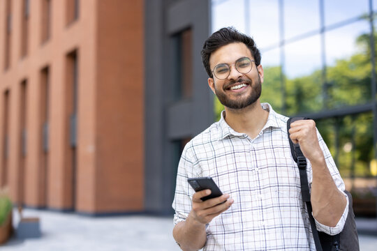 Fototapeta A smiling man holds a phone in front of a modern building.
