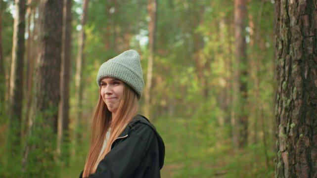 Young girl wearing gray beanie and black jacket smiling while playfully holding tent pole during camping setup in forest as sunlight filters through trees and friend stands in background