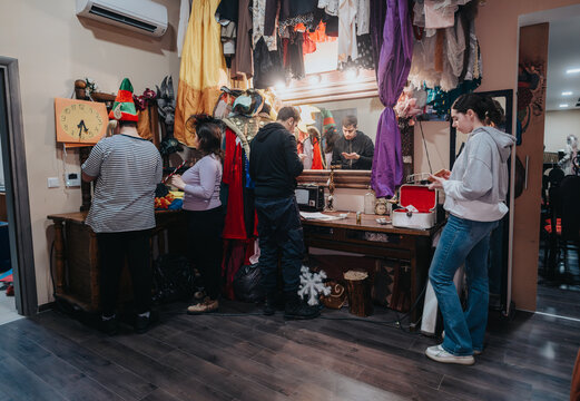 Actors and crew members organize costumes and apply makeup in a vibrant theater dressing room, surrounded by accessories and props, as they prepare backstage for a live performance.