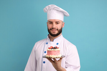 Happy confectioner in uniform holding delicious cake with berries on light blue background