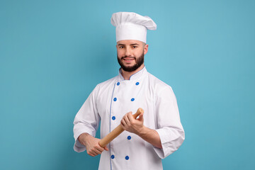 Happy confectioner in uniform holding rolling pin on light blue background