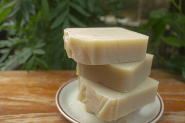 Handmade Centella Herbal Soap Bars. A stack of natural handmade soap bars infused with Centella asiatica (Gotu Kola), placed on a white dish over a wooden table with green garden background.