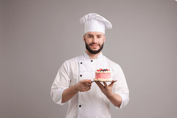 Happy confectioner in uniform holding delicious cake with berries on light grey background
