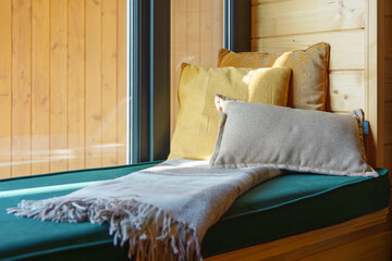 Comfortable seating nook features soft cushions and warm blanket, illuminated by natural light. The wooden walls create rustic yet inviting atmosphere perfect for relaxation. Home interior detail