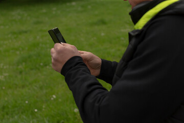 An individual is seen operating a mobile phone while standing on a patch of grass, dressed in a dark jacket that features a reflective strip for visibility in low light conditions