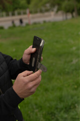 An individual is utilizing a smartphone as they hold a small tool in a lush outdoor park setting filled with vibrant green grass. This combines elements of modern technology with nature