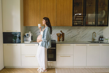 A pregnant woman stands in a stylish kitchen, holding a cup tea and reflecting on impending...