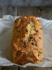 Close-up of a banana bread with chocolate chips on baking paper on a rustic wooden table. Natural light. Breakfast concept, artisanal pastries, home cooking.