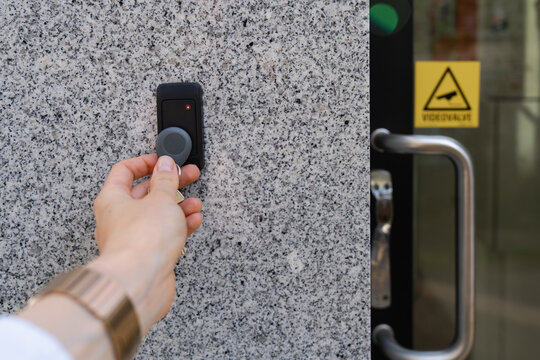 Close-up of a female hand holding a keychain with a key fob near a key reader access device on a building&rsquo;s exterior wall with door and video surveillance sign. Secure access control technology