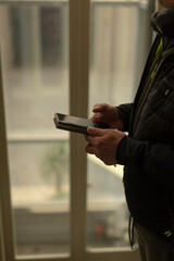 A person is indoors, holding a smartphone while gazing outside through a window, vividly...