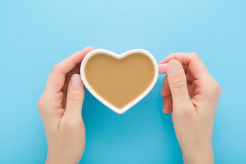 Young adult woman hands holding white heart shape cup of fresh coffee with milk on light blue table background. Pastel color. Closeup. Point of view shot. Top down view.