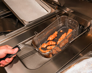 Freshly fried chicken strips in a deep fryer basket, lifted from hot oil in a commercial kitchen. Stainless steel equipment and crispy food preparation in focus.