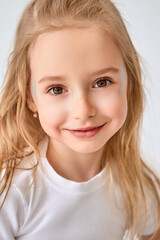Close-up portrait of a lovely little girl with long blond hair, wearing a white T-shirt. Blue glitter sparkles on her cheeks. The child smiles, creating a friendly and joyful impression.