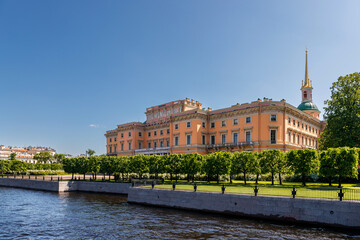 Mikhailovsky Castle with a golden spire stands by the river, with even trees growing along the bank.