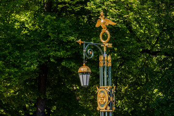A decorative street lamp with a golden double-headed eagle and a lantern, against a background of green tree foliage.