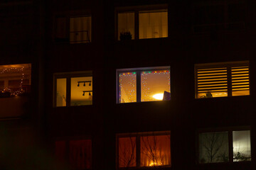 In a dim apartment building, glowing windows with holiday lights create a cozy atmosphere, casting enchanting shadows that foster a warm and inviting ambiance this evening